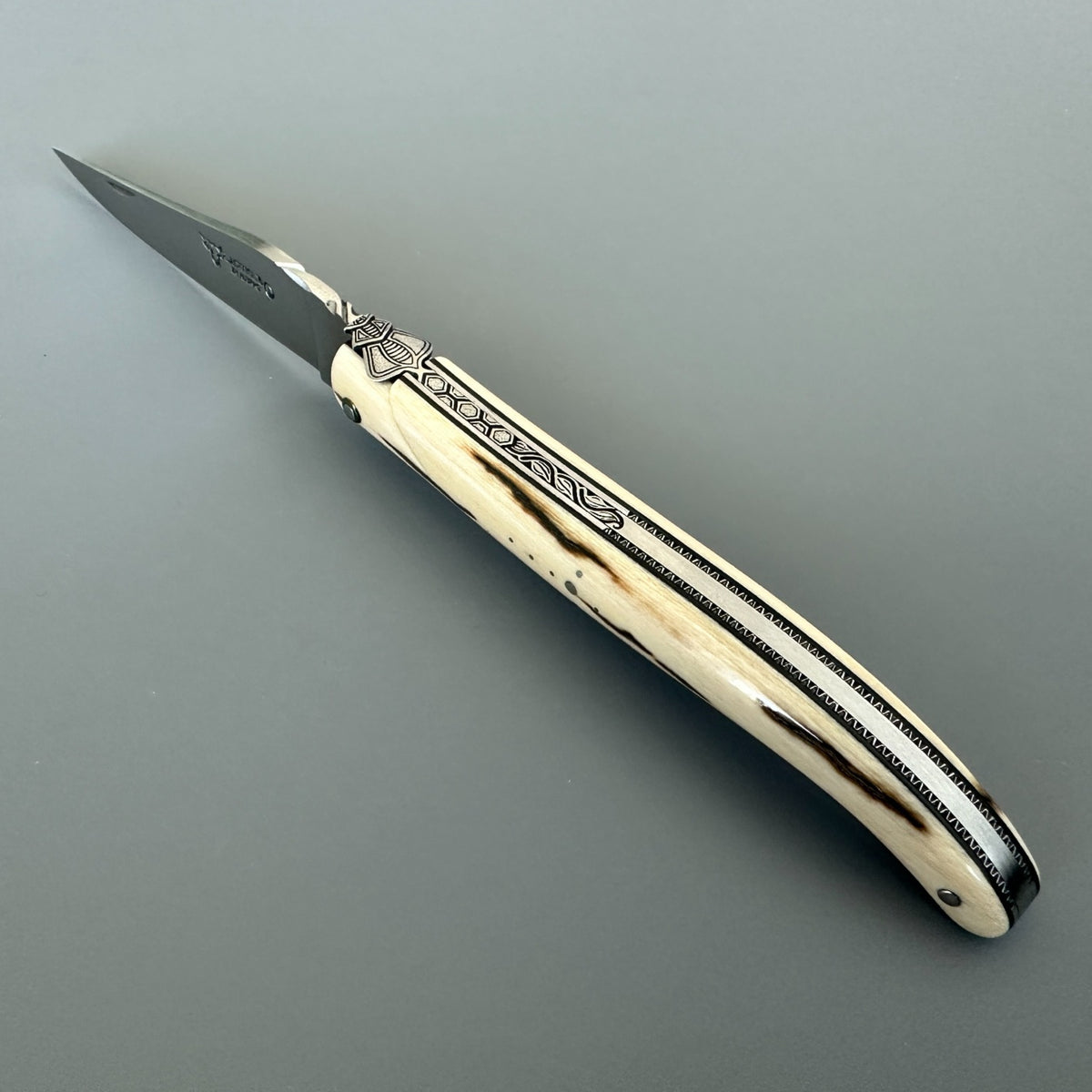 Open Laguiole en Aubrac mammoth ivory folding knife with dark handle streaks on a gray background.