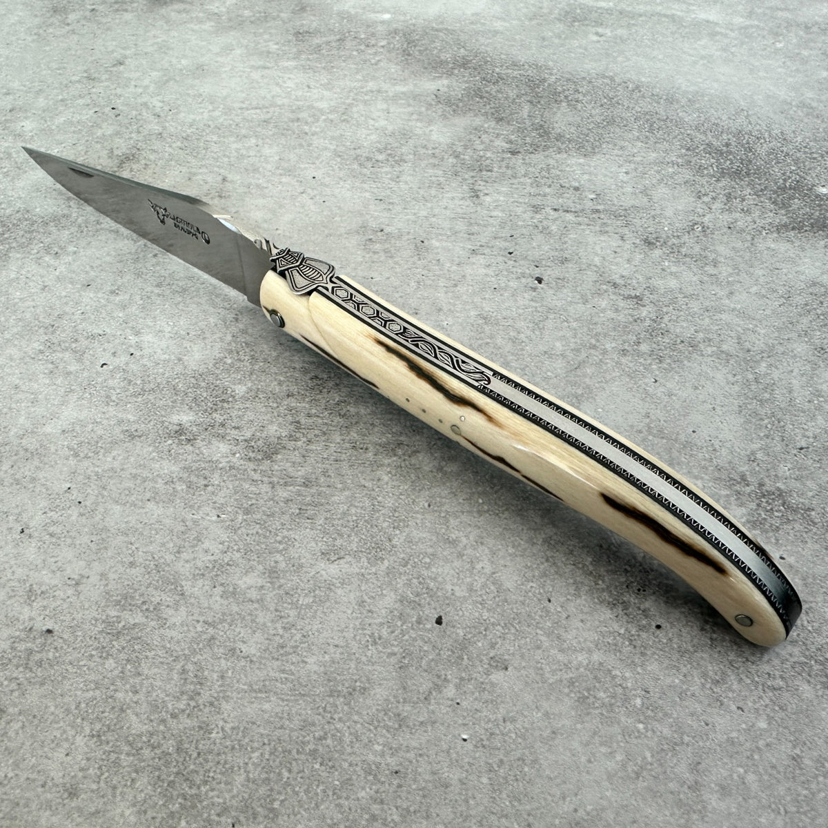 Open Laguiole en Aubrac mammoth ivory folding knife with dark handle streaks on a textured gray background.