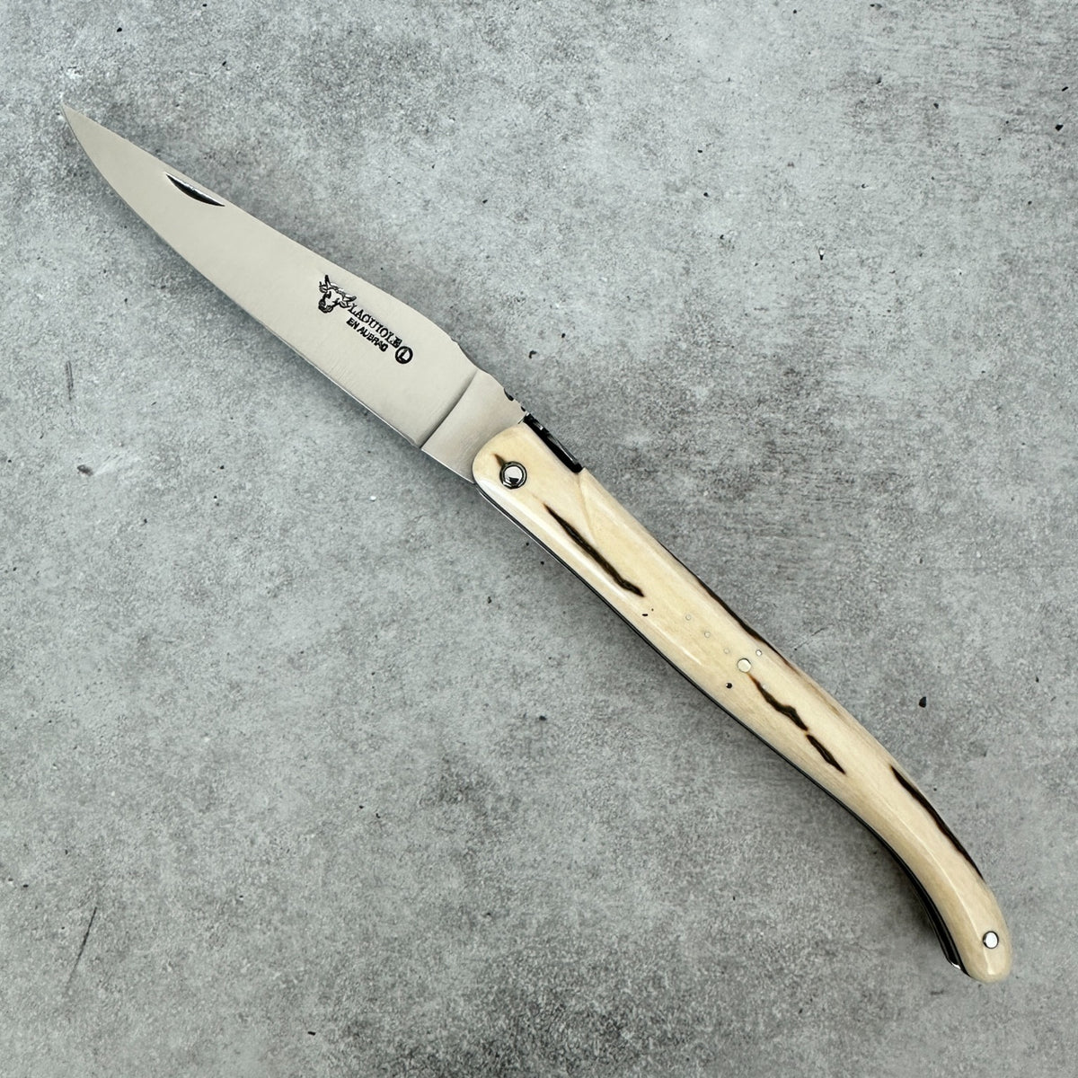 Angled Laguiole en Aubrac mammoth ivory folding knife showing its open blade on a textured background.
