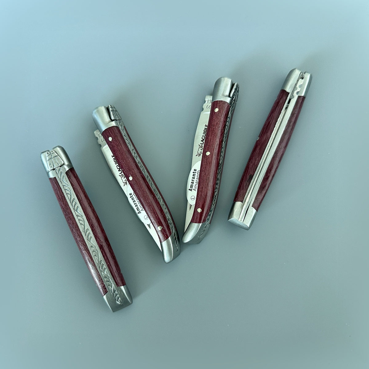 Four Laguiole en Aubrac Mini Folding Knives with Amaranth handles arranged on a natural wood surface.
