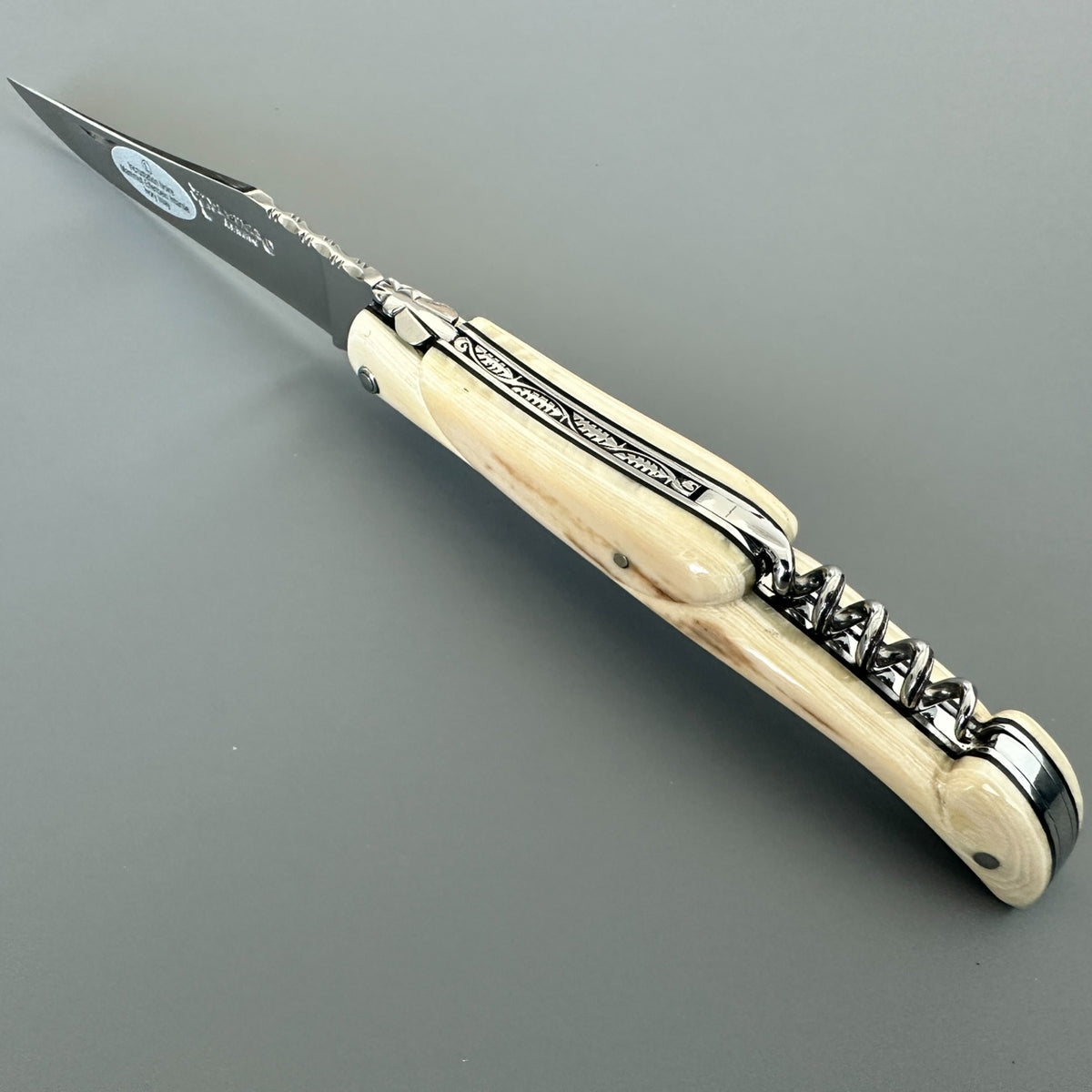 Angled view of an open Laguiole en Aubrac folding knife with a pale handle, engraved spring, and corkscrew.