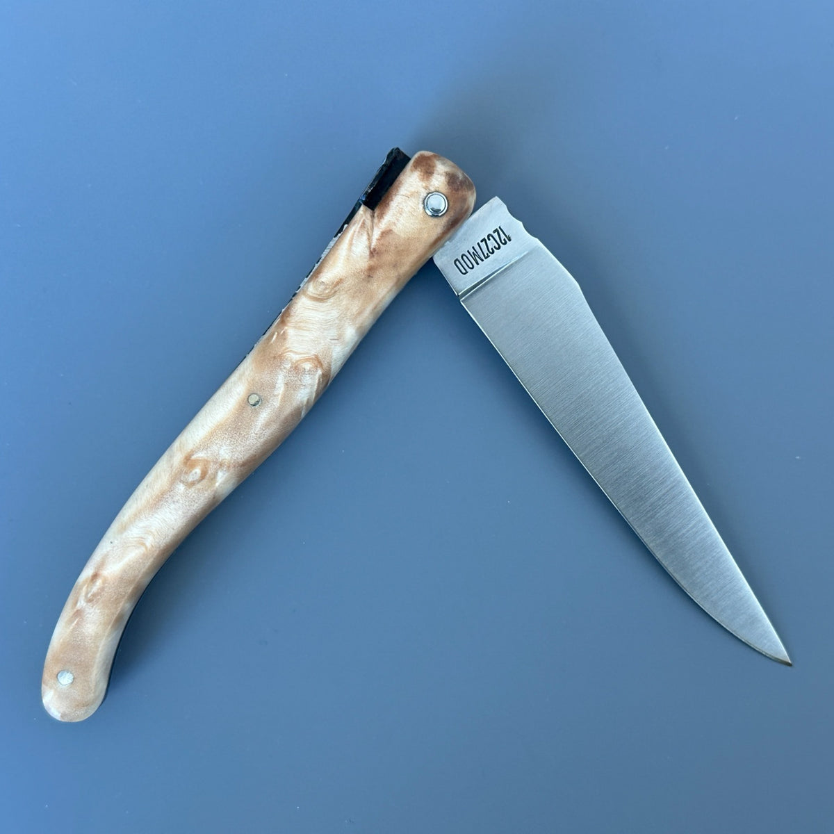 Reverse side of a partially open Laguiole en Aubrac folding knife showing the figured poplar handle and satin blade.