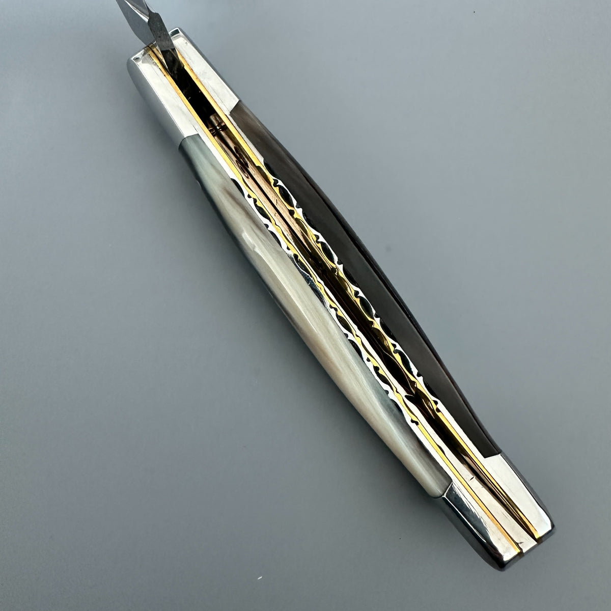 Laguiole en Aubrac double-plated pocket knife with a solid horn handle and polished silver-toned metal blades and brass inner spine, shown open on a neutral gray background.