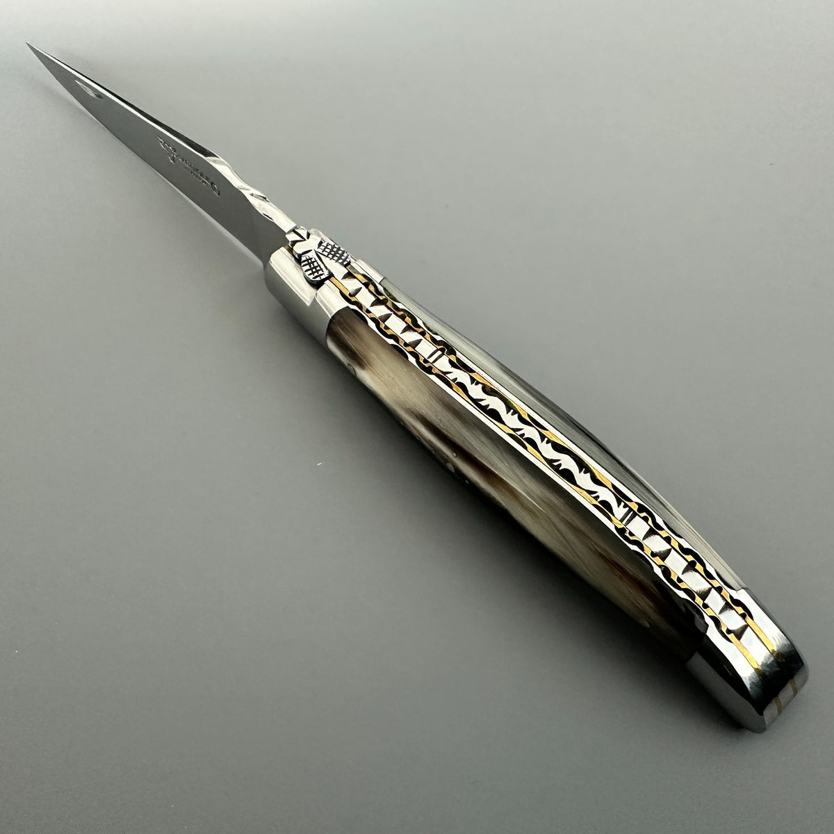 Laguiole en Aubrac folding knife with a solid horn handle featuring intricate brass and silver filigree inlays, and a silver stainless steel blade, shown on a neutral gray background.