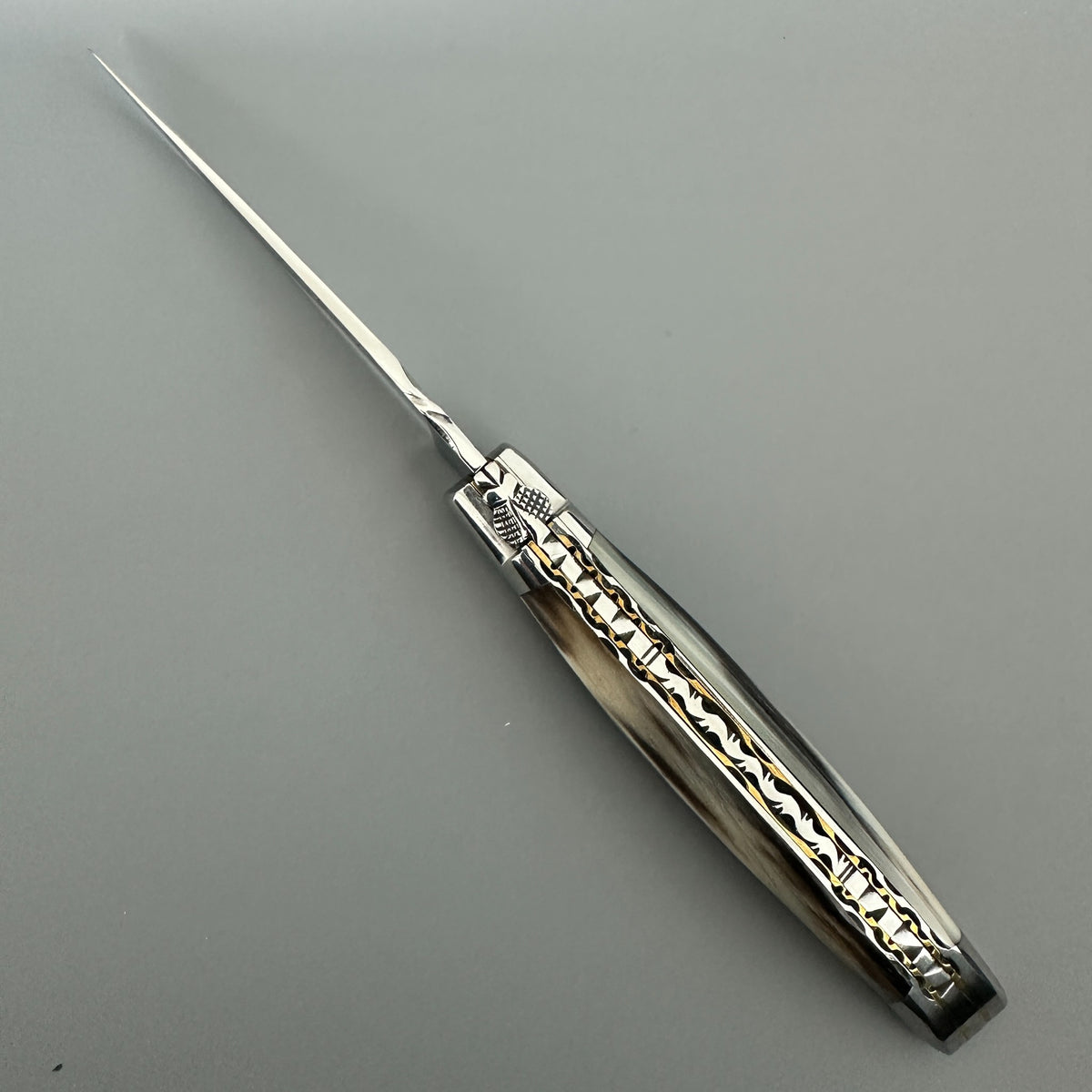 Cenital view of the Laguiole en Aubrac double-plated pocket knife with a solid horn handle, featuring intricate gold-toned floral engravings on the metal collar and handle.