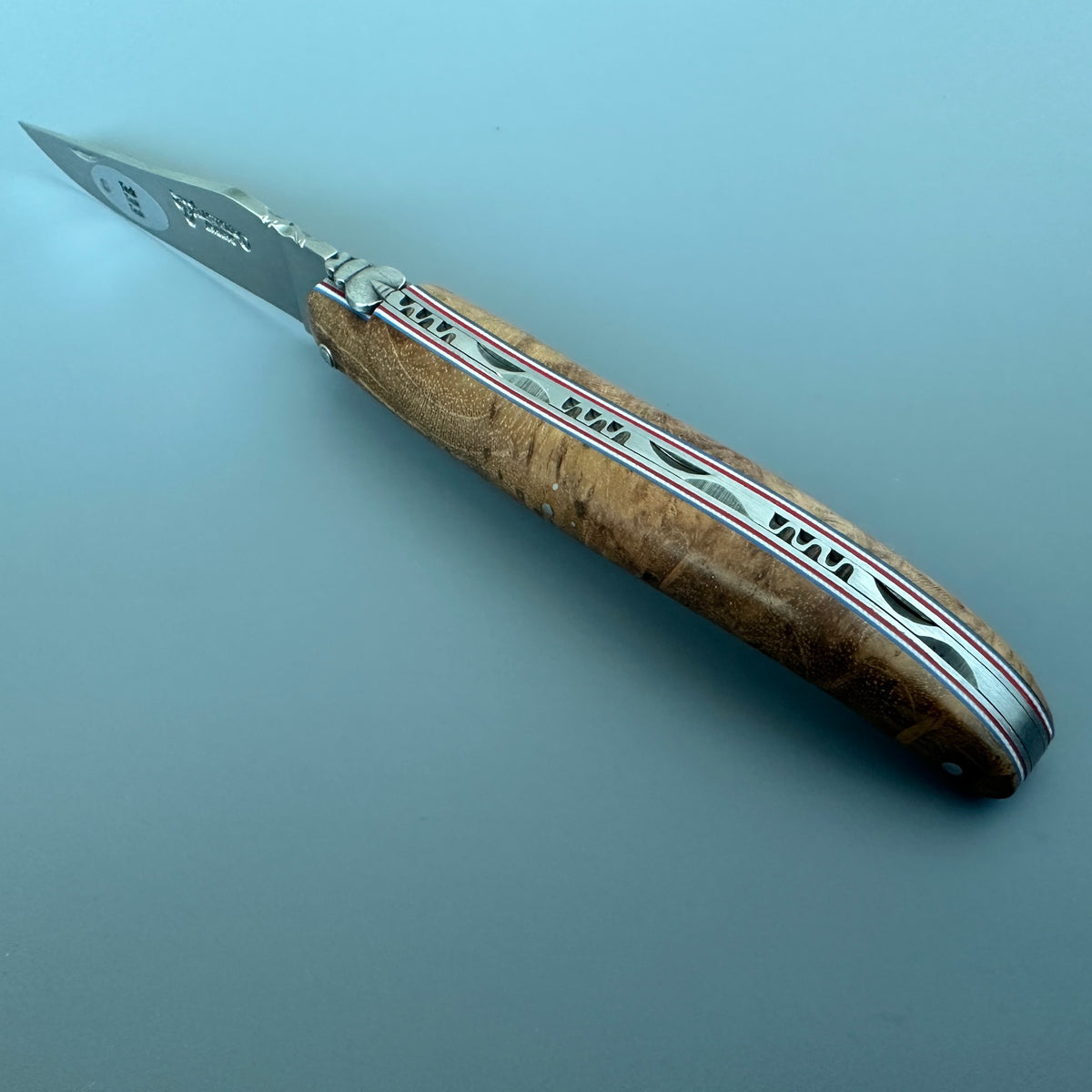 A folding pocket knife with a satin silver blade and a custom wooden handle featuring inlaid silver, red, and blue stripes, shown on a solid blue-gray background.