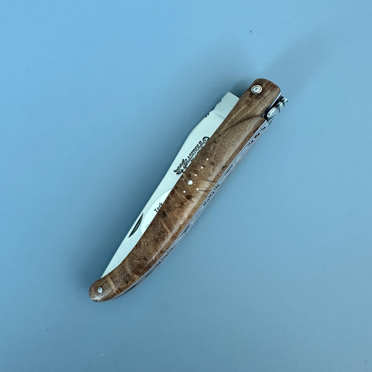 A Laguiole en Aubrac folding pocket knife with a brown wood-grain handle and a silver metal blade, shown open on a solid light blue background. 