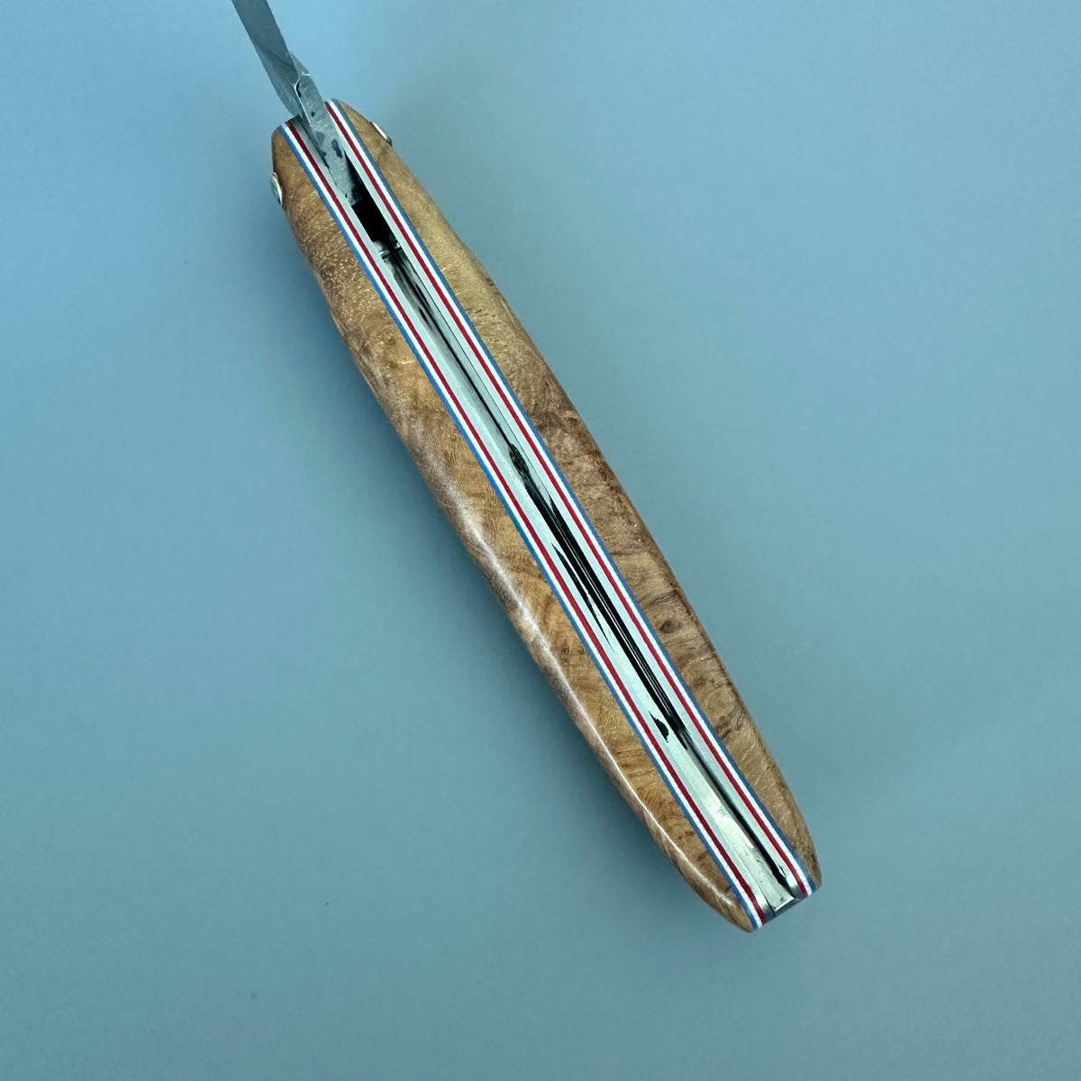A folding pocket inner spine knife with a polished wood handle and a silver metal blade, featuring a red, white, and blue striped inlay along the center, set against a solid light blue background.