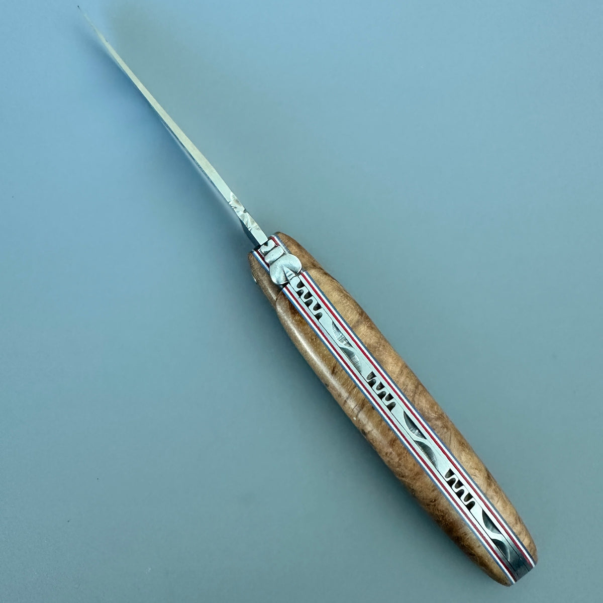 Cenital view folding pocket knife with a polished silver blade and a decorative handle made of light brown wood and silver metal inlay, shown on a solid blue-gray background.