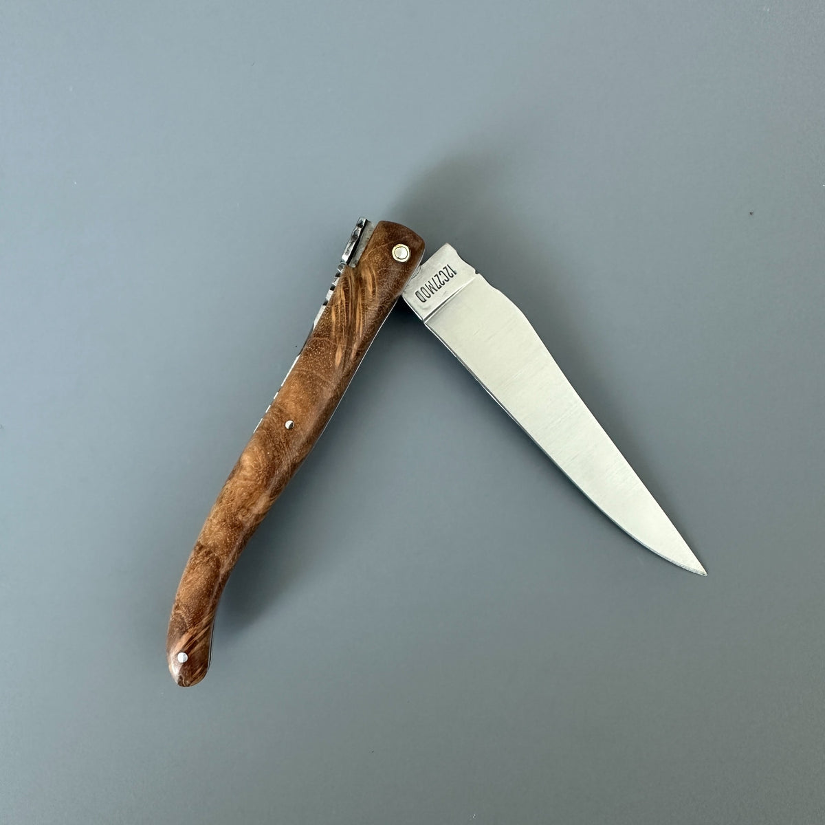 Reverside side of a folding pocket knife with a satin finish silver blade and a textured brown teak wood handle, shown open on a solid gray background. 